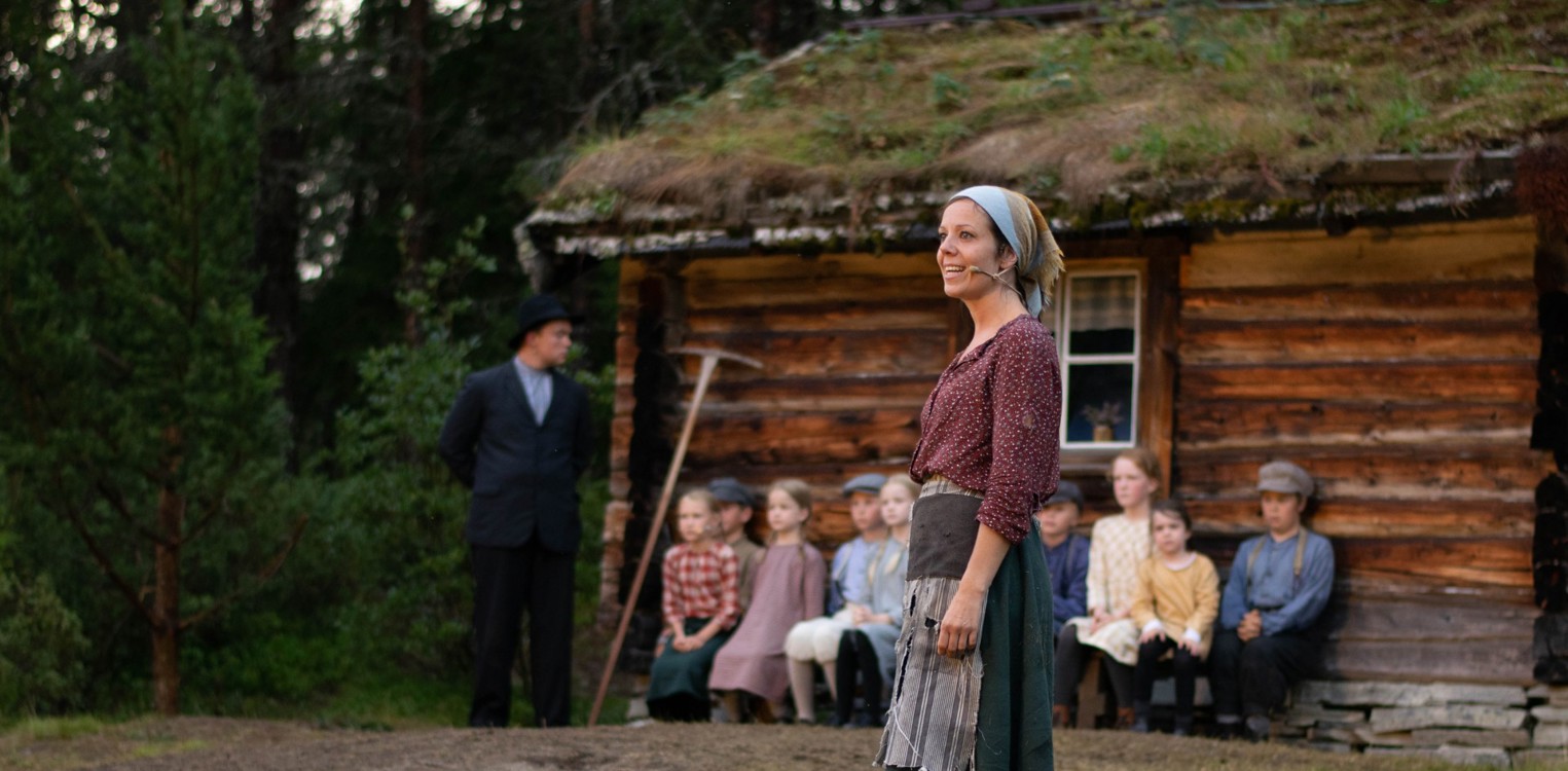 En kvinne i gamle klær står foran en torvtakhytte, med en mann og flere barn i historiske klær sittende langs hytteveggen bak henne.