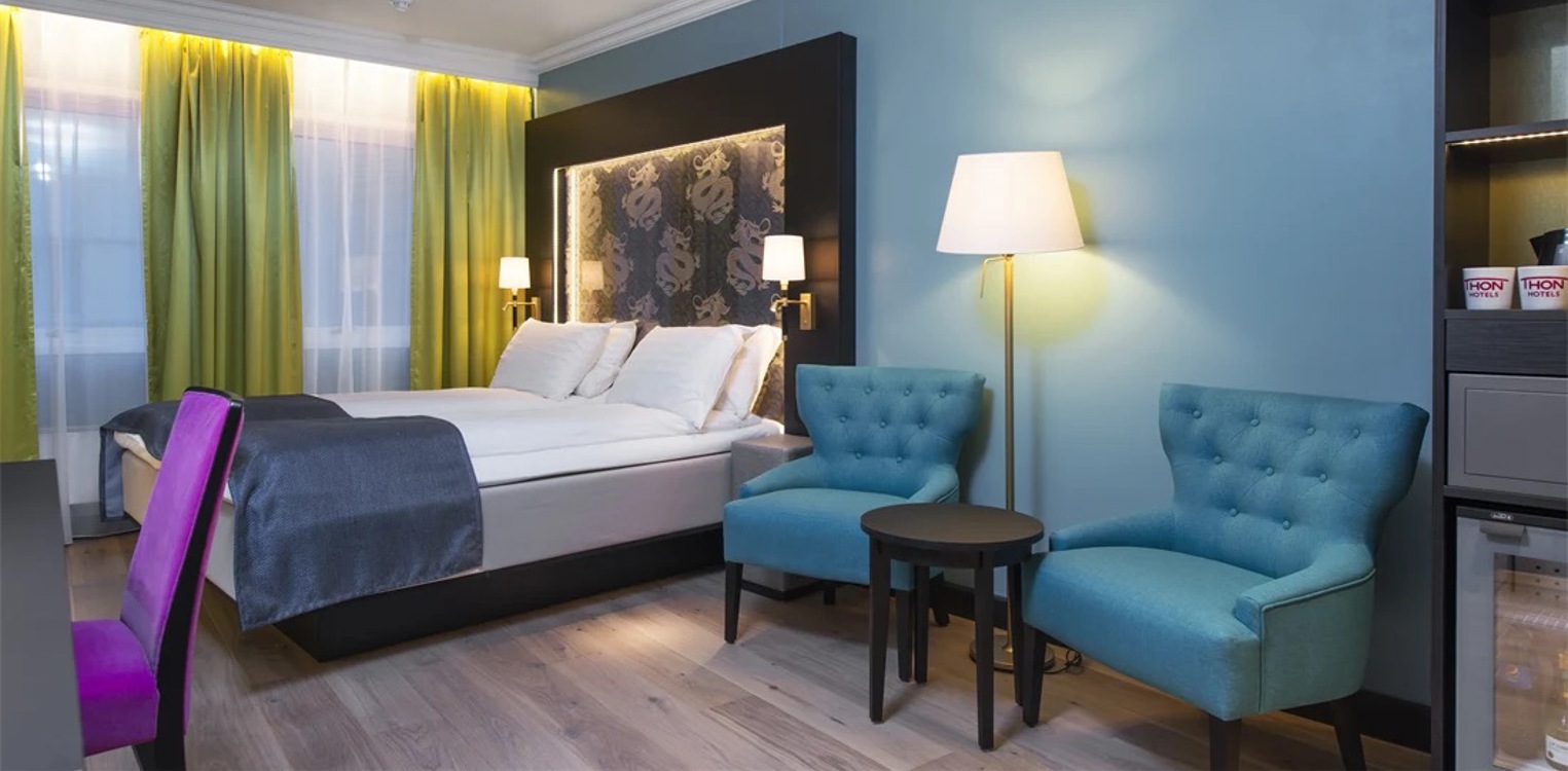 Hotellrom med to stoler og et bord, stålampe bak, og en seng ved vinduer med grønne gardiner.