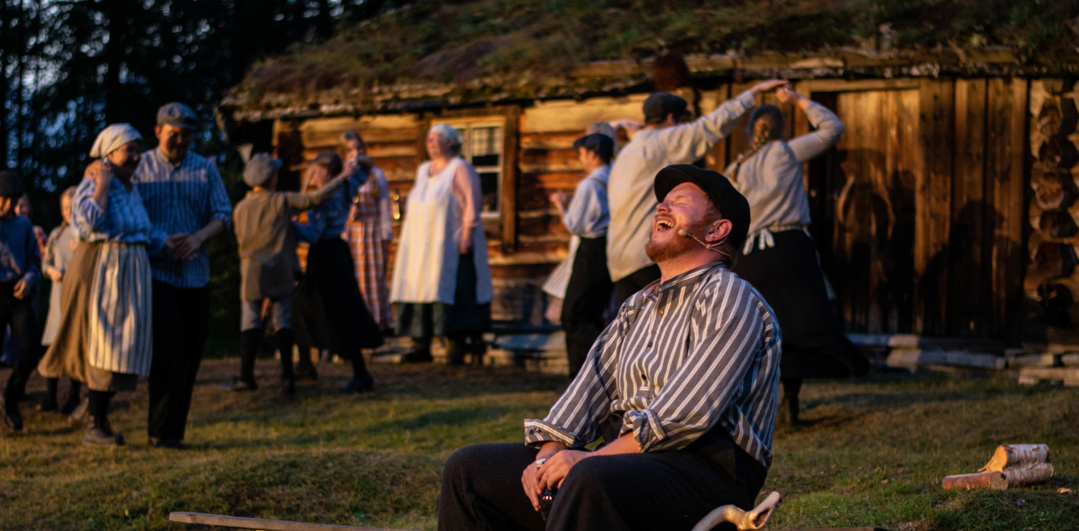 Skuespillere i historiske klær foran hytte med torvtak; en mann sitter på en benk i forgrunnen og ler mens andre danser bak ham.