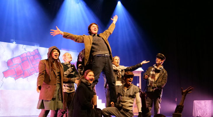 Ensemble av unge på scene i historiske klær; en mann står med åpne armer oppå lårene til to som kneler i en avslutningsposisjon.