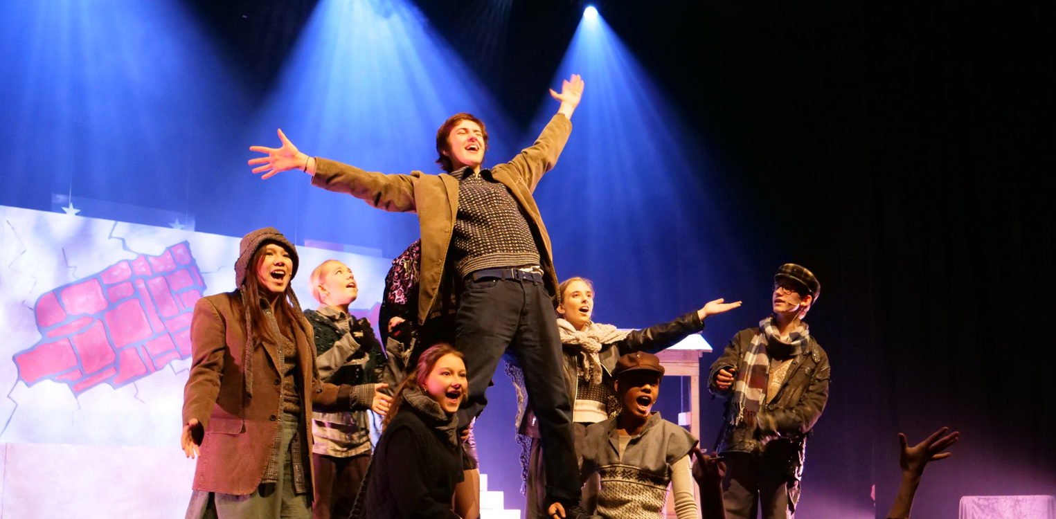 Ensemble av unge på scene i historiske klær; en mann står med åpne armer oppå lårene til to som kneler i en avslutningsposisjon.