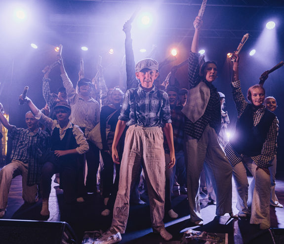 Ung gutt står i forgrunnen på SplæshCamp-scenen, med ungdommer bak seg som holder aviser i været under blått og gult scenelys.