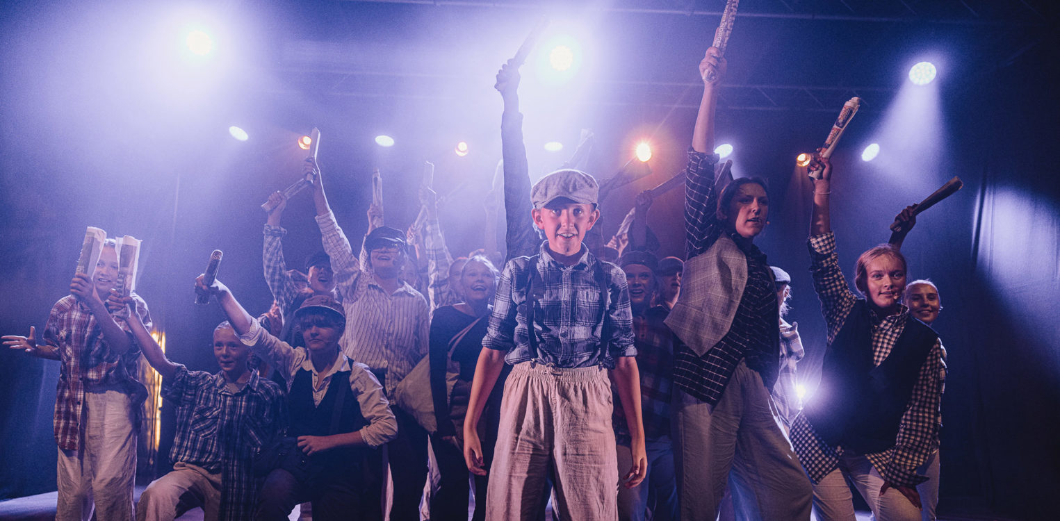 Ung gutt står i forgrunnen på SplæshCamp-scenen, med ungdommer bak seg som holder aviser i været under blått og gult scenelys.