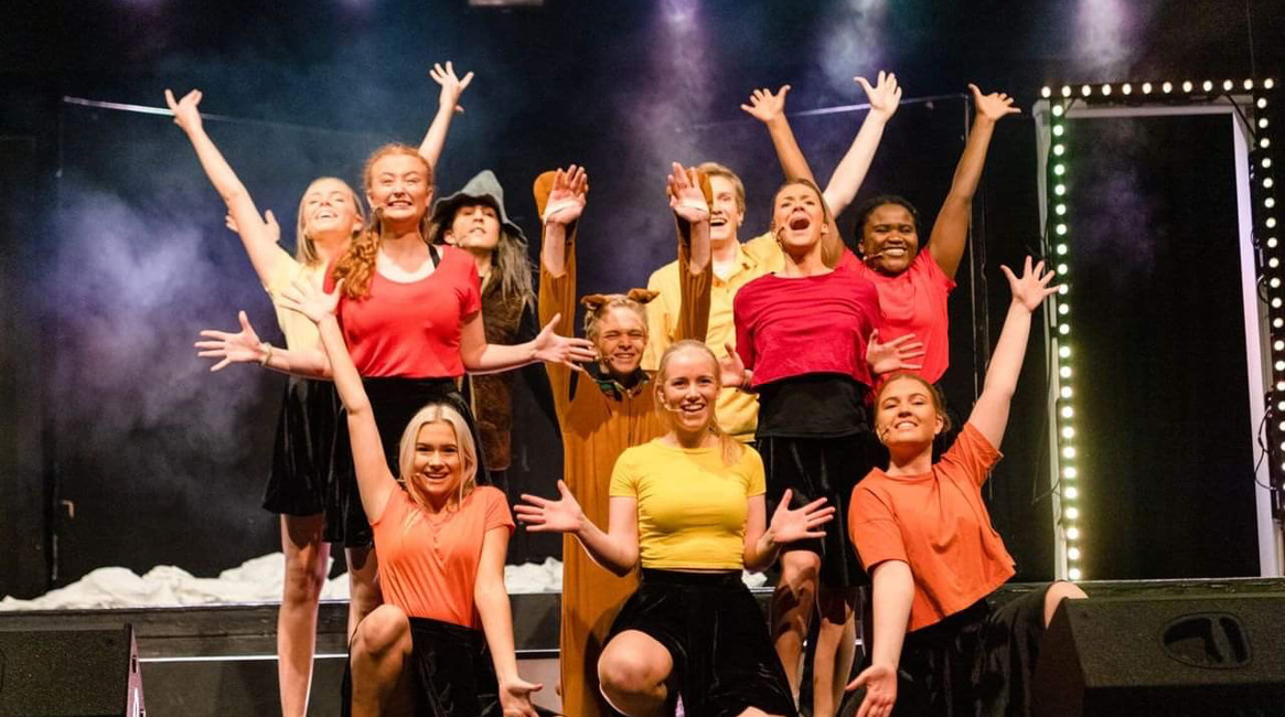 Skuespillere i oransje, gule og røde overdeler står i avslutningspositur med en kattfigur i midten.