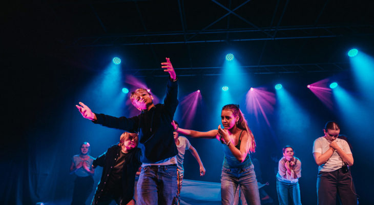 Ungdommer opptrer på en scene med blått og rosa lys; en gutt står i front med armene åpne mens en jente holder en hånd under hans venstre arm.