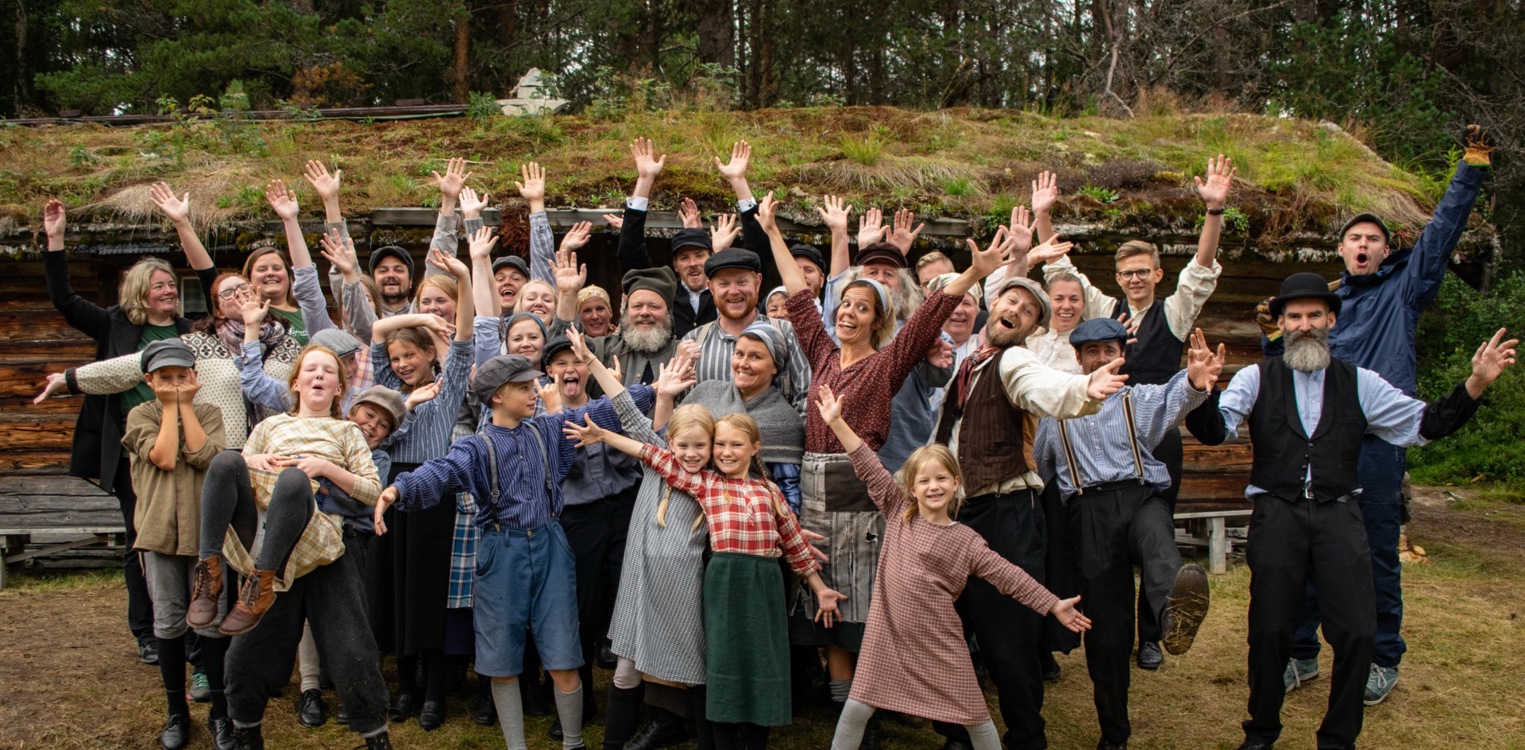 Ensemble fra utendørs teater foran en torvtak-hytte, kledd i historiske klær.
