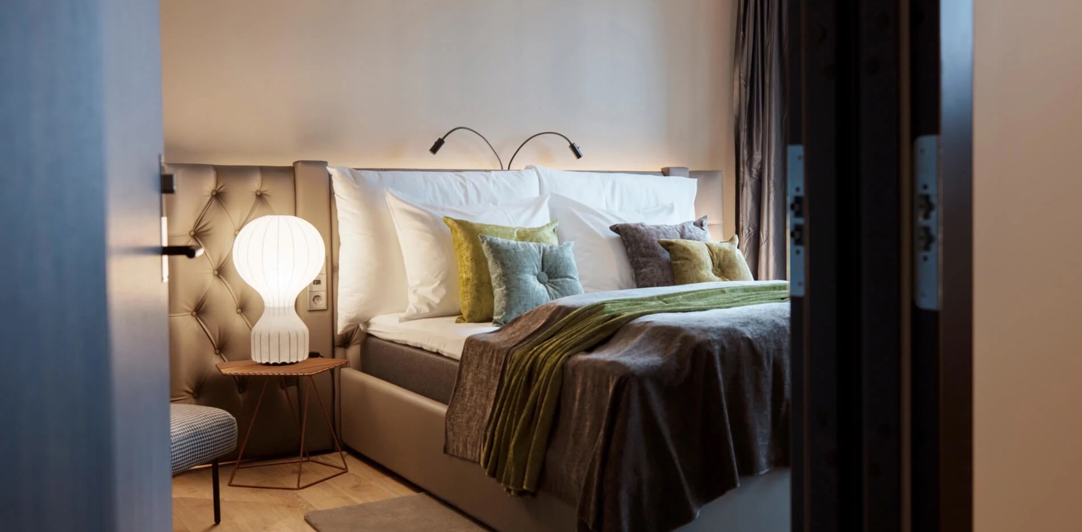 Hotellrom sett fra døren, med oppredd seng, pynteputer og nattbord med lys.