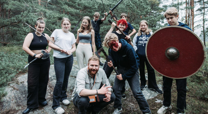 Ungdommer i stagefightkurs med sverd, øks og skjold, en med fake blod i ansiktet; instruktør sitter på huk foran dem i skogmiljø.