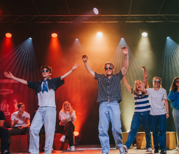 Ungdommer på scene med solbriller; to gutter i front klapper over hodet mens tre jenter står bak dem.